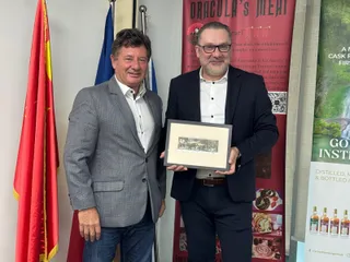 Doi bărbați stau în picioare lângă un steag și un panou publicitar, unul dintre ei ținând un cadou încadrat.