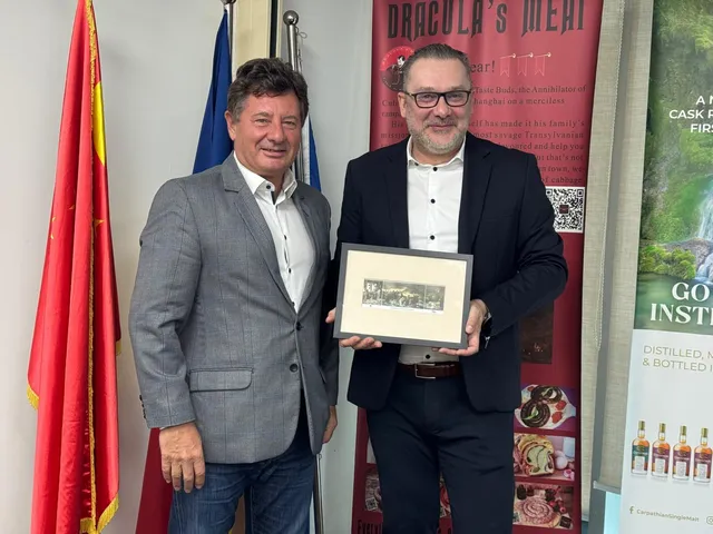 Doi bărbați stau în picioare lângă un steag și un panou publicitar, unul dintre ei ținând un cadou încadrat.