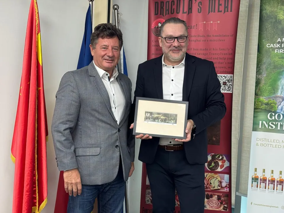 Doi bărbați stau în picioare lângă un steag și un panou publicitar, unul dintre ei ținând un cadou încadrat.