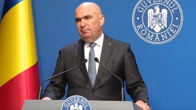 Un bărbat cu părul scurt și costum închis stă în spatele unui podium cu microfoane în fața unui fundal albastru cu un emblema circulară.