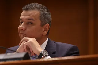 Un bărbat cu părul scurt și grij, purtând un sacou închis la culoare și o cămașă albă, stă cu mâinile sub bărbie.