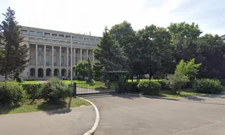O clădire gri cu coloane înalte este vizibilă în spatele unor copaci verzi și a unui gard metalic negru.