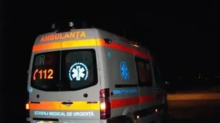 O ambulanță albă cu benzi reflectorizante portocalii și inscripții galbene este parcată pe un drum în timpul nopții.