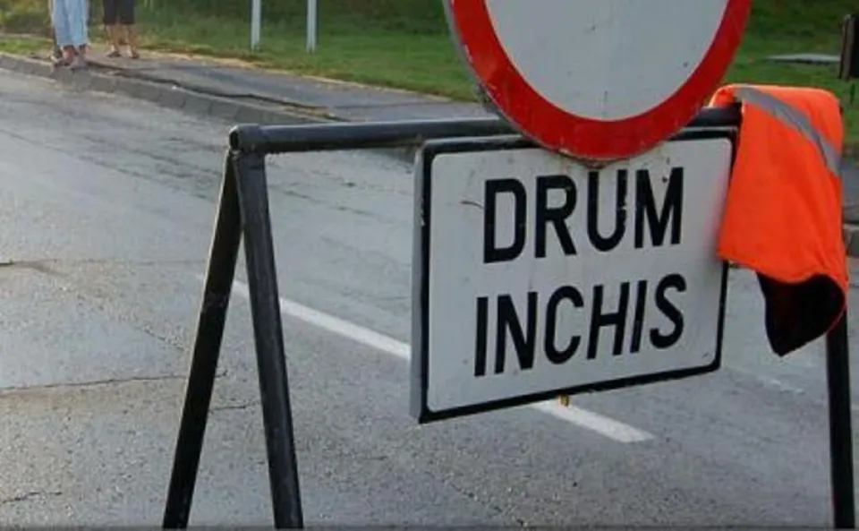 Un semn de circulație alb cu margini negre pe care scrie drum închis este montat pe un suport metalic pe un drum asfaltat.
