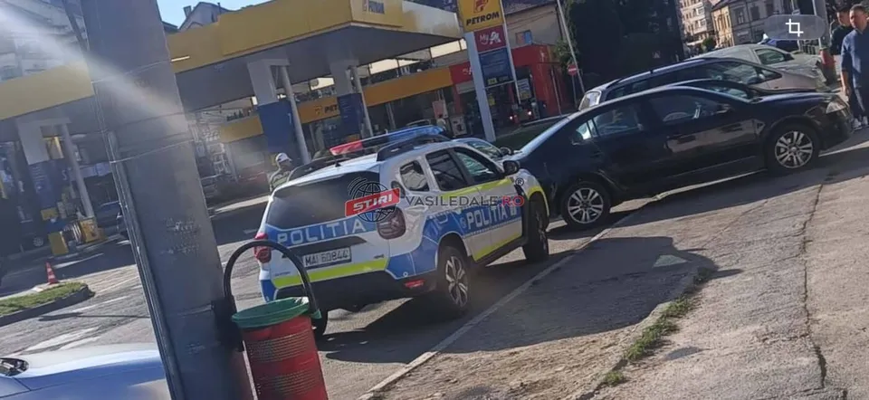 O mașină de poliție albastră și albă este parcată pe un drum lângă un vehicul negru și o benzinărie sub lumina soarelui.