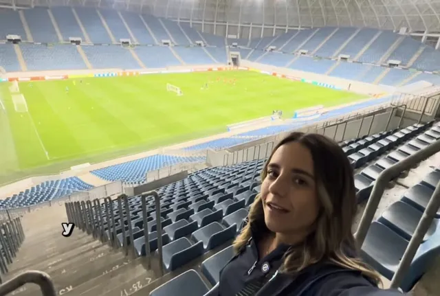 O femeie cu părul lung și deschis la culoare stă în tribunele unui stadion, având în fundal un teren de fotbal verde și scaune albastre.