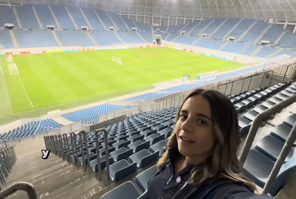O femeie cu părul lung și deschis la culoare stă în tribunele unui stadion, având în fundal un teren de fotbal verde și scaune albastre.