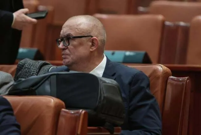 Un bărbat cu ochelari și costum închis se așază pe un scaun de piele maro într-o încăpere cu scaune similare în fundal.