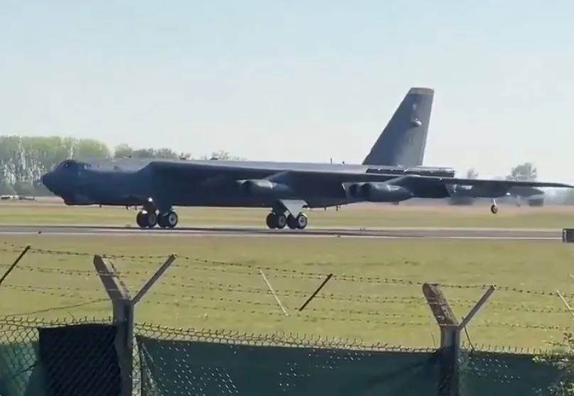 Un avion militar de mari dimensiuni cu fuselaj gri este poziționat pe o pistă de aterizare lângă un teren cu iarbă și un gard de sârmă ghimpată.