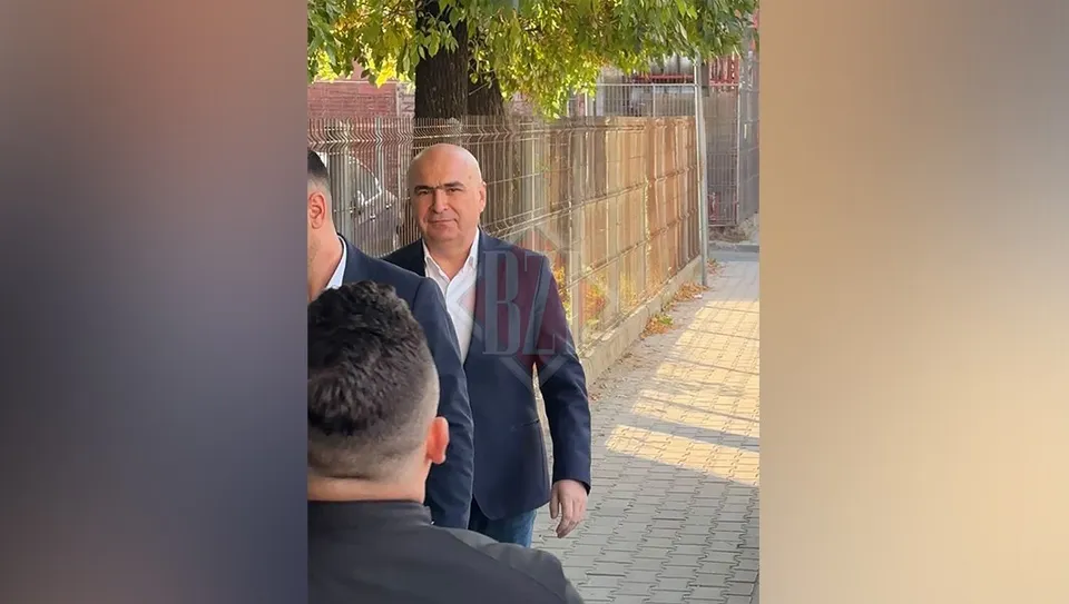 Un bărbat cu părul scurt și costum închis merge pe un trotuar lângă un gard metalic și un copac.