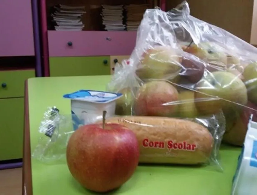 Un măr roșu, un produs de panificație cu textul Corn Scolar și un recipient de iaurt sunt așezate pe o masă verde.