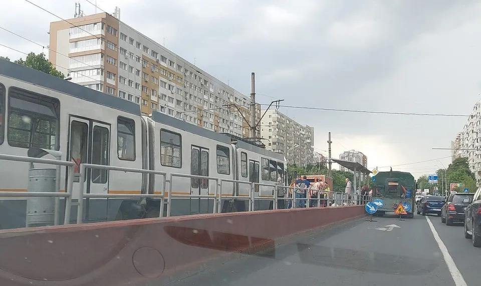 Un tramvai alb și gri circulă pe o stradă lângă un bloc înalt și vehicule parcate.