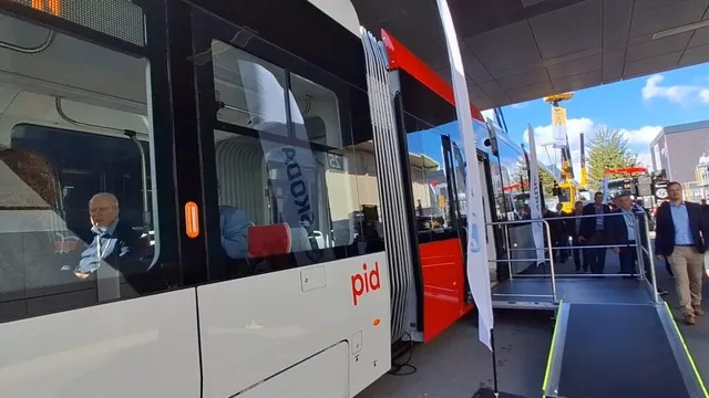 Un tramvai alb cu detalii roșii este parcat lângă o rampă de acces, în timp ce câțiva bărbați stau în apropiere sub un acoperiș.