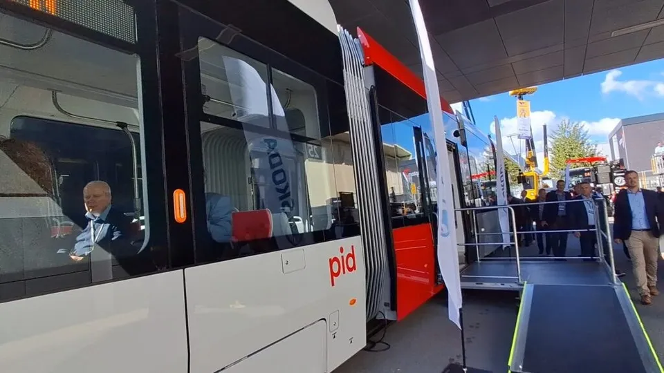 Un tramvai alb cu detalii roșii este parcat lângă o rampă de acces, în timp ce câțiva bărbați stau în apropiere sub un acoperiș.