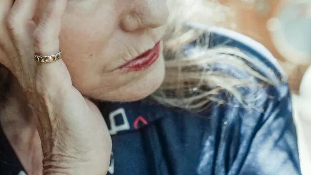O persoană vârstnică cu părul alb își sprijină capul pe o mână, purtând un inel subțire și o cămașă închisă la culoare.