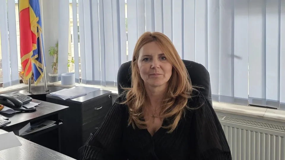 O femeie cu părul blond și bluză neagră stă la un birou într-un birou cu jaluzele verticale și un steag în fundal.