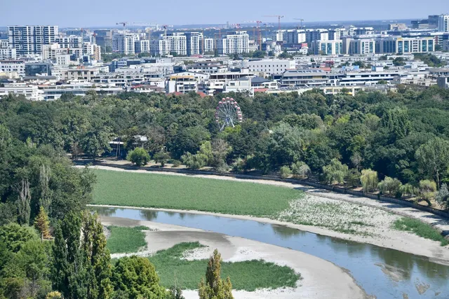 O vedere panoramică asupra unui parc verde cu un lac îngust, un iaz de nisip și o roată romantică în depărtare sub un cer senin.