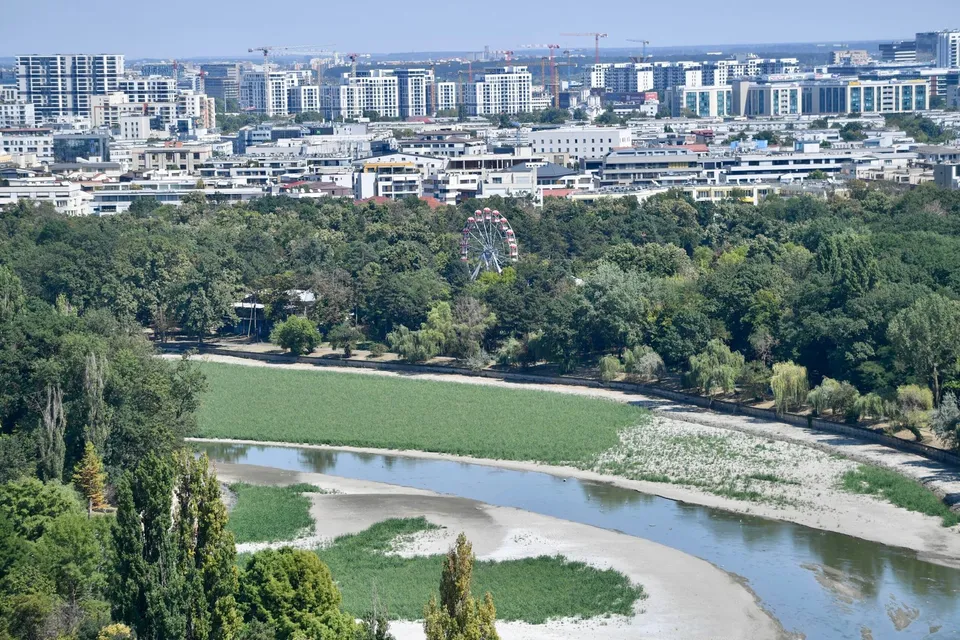 O vedere panoramică asupra unui parc verde cu un lac îngust, un iaz de nisip și o roată romantică în depărtare sub un cer senin.