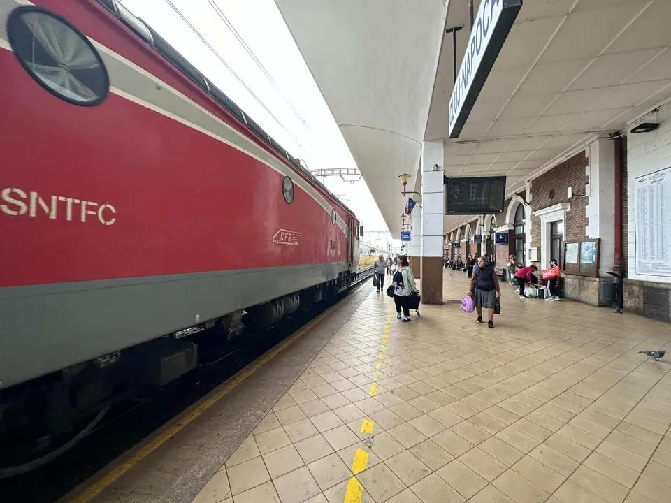 Un tren roșu și gri stă pe un binar lângă un peron de gară unde câțiva oameni merg sau stau pe scaune.