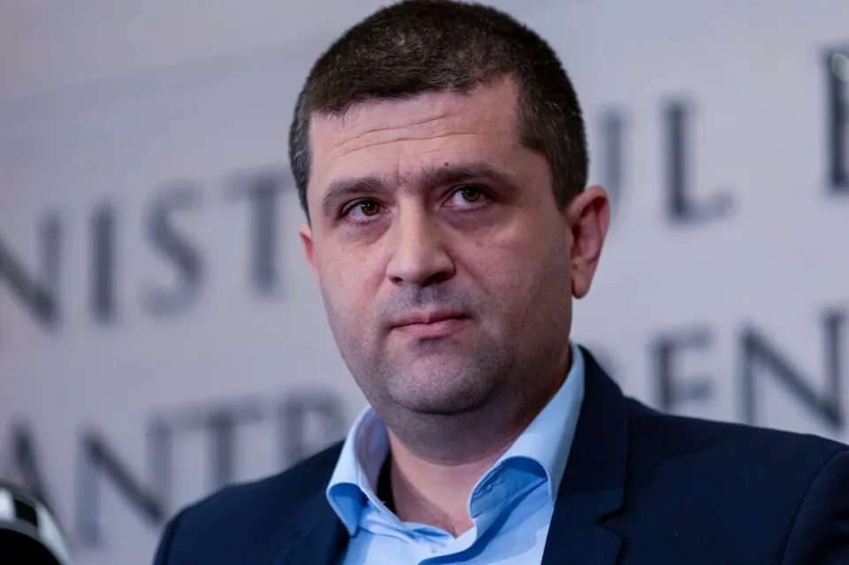 Un bărbat cu păr scurt și cămașă albastră poartă un sacou în fața unui fundal cu litere întunecate.