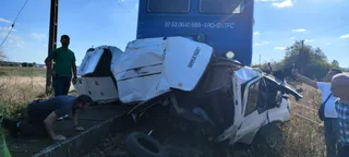 O autoutilitară albă este zdrobită pe o cale ferată lângă un tren albastru, în timp ce trei bărbați privesc vehiculul distrus.