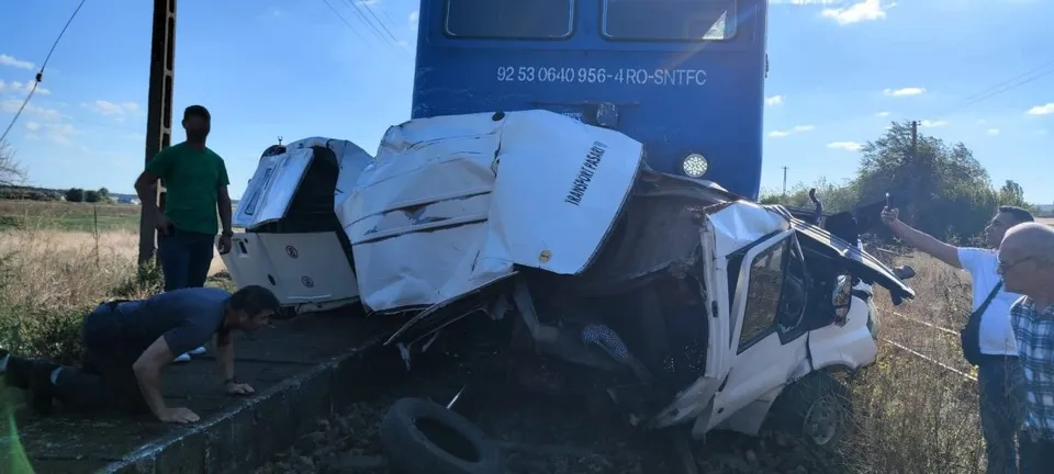 O autoutilitară albă este zdrobită pe o cale ferată lângă un tren albastru, în timp ce trei bărbați privesc vehiculul distrus.