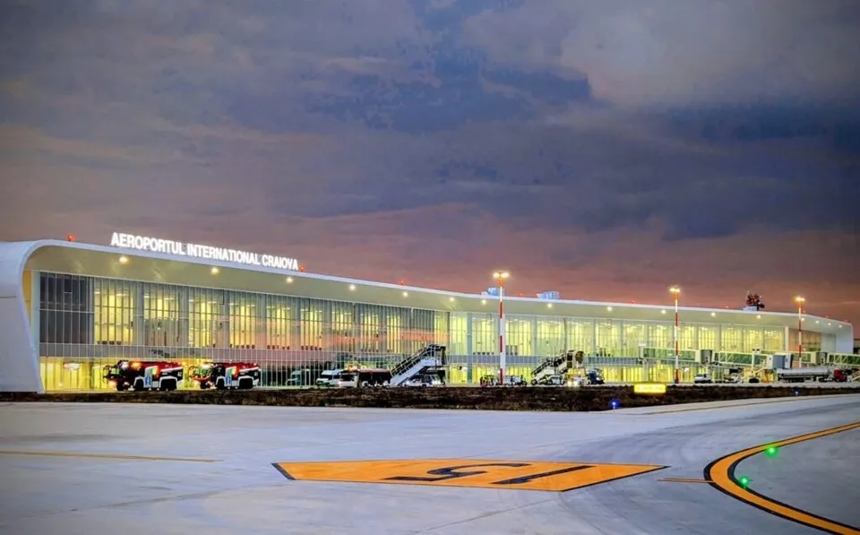 O clădire de terminal de aeroport cu ferestre mari iluminate este situată lângă o pistă de asfalat sub un cer de seară.