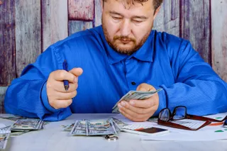 Un bărbat cu cămașă albastră stă la o masă cu bancnote, un calculator și ochelari în fața sa.
