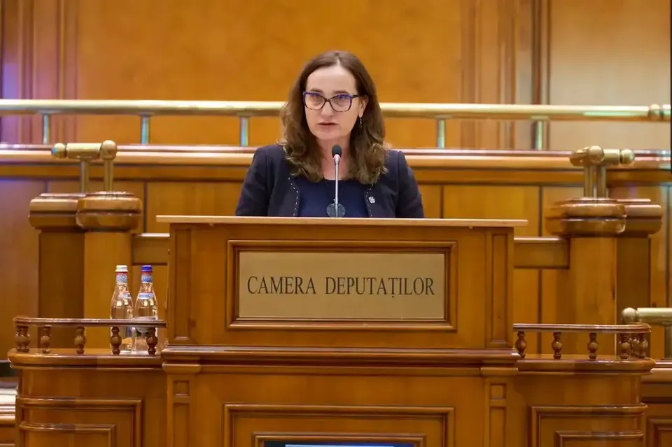 O femeie cu ochelari și părul castaniu stă în spatele unui podium de lemn pe care este scris textul CAMERA DEPUTAȚILOR.