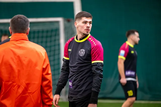 Un bărbat cu o cămașă neagră cu mâneci lungi și detalii roz se află pe un teren de fotbal lângă un alt bărbat în echipament sportiv.
