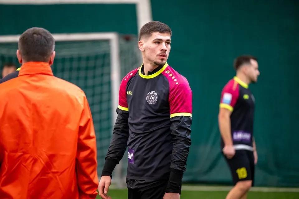 Un bărbat cu o cămașă neagră cu mâneci lungi și detalii roz se află pe un teren de fotbal lângă un alt bărbat în echipament sportiv.