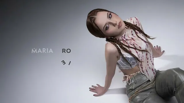 O femeie cu părul împletit poartă un tricou tip crop top și pantaloni de piele, stând într-o poziție înclinată pe un fundal gri.