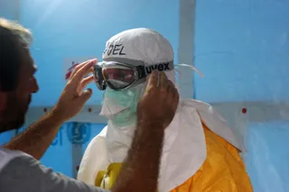Un bărbat îmbrăcat într-un costum de protecție alb, cu mască și ochelari de protecție, este ajutat să își ajusteze echipamentul de către o altă persoană.