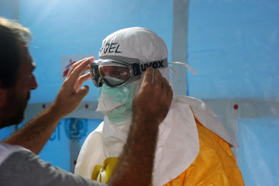 Un bărbat îmbrăcat într-un costum de protecție alb, cu mască și ochelari de protecție, este ajutat să își ajusteze echipamentul de către o altă persoană.