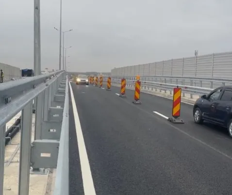 O mașină neagră circulă pe un drum asfaltat lângă bariere metalice și conuri de trafic portocalii cu dungi galbene sub un cer înnorat.