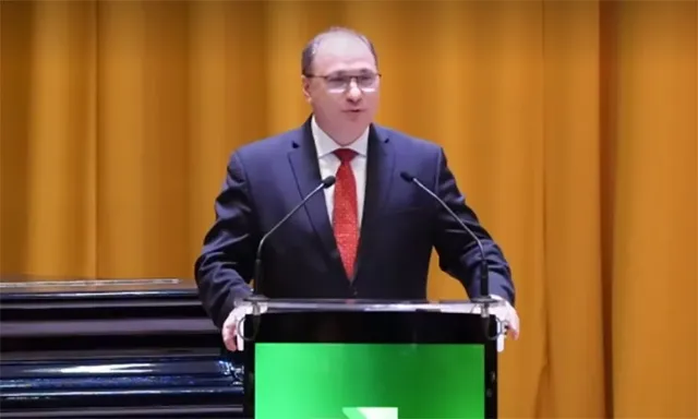 Un bărbat cu ochelari și costum închis stă în spatele unui podium negru cu un panou verde în fața unui fundal de draperii galbene.