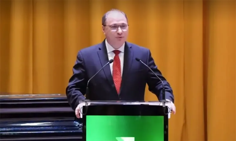 Un bărbat cu ochelari și costum închis stă în spatele unui podium negru cu un panou verde în fața unui fundal de draperii galbene.