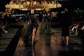 O femeie poartă o rochie neagră lungă pe o pistă de catwalk sub un candelabru mare iluminat.