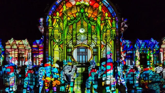 O clădire este acoperită de proiecții colorate cu modele geometrice și vegetale, în timp ce siluete de oameni sunt vizibile în prim-plan.