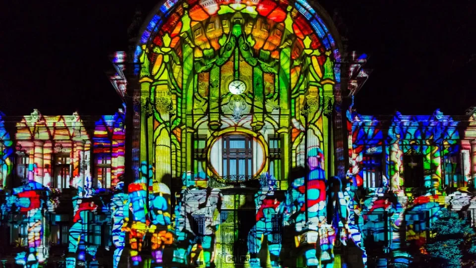 O clădire este acoperită de proiecții colorate cu modele geometrice și vegetale, în timp ce siluete de oameni sunt vizibile în prim-plan.