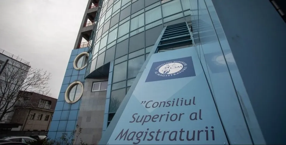 O clădire modernă cu fațadă de sticlă albastră prezintă un panou alb pe care scrie textul în limba română despre Consiliul Superior al Magistraturii.