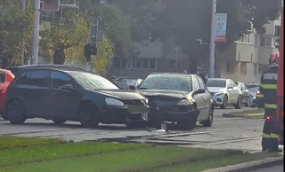 Două autoturisme de culoare închisă sunt oprite pe o stradă lângă o zonă cu iarbă, în prezența unui membru al corpului de pompieri.