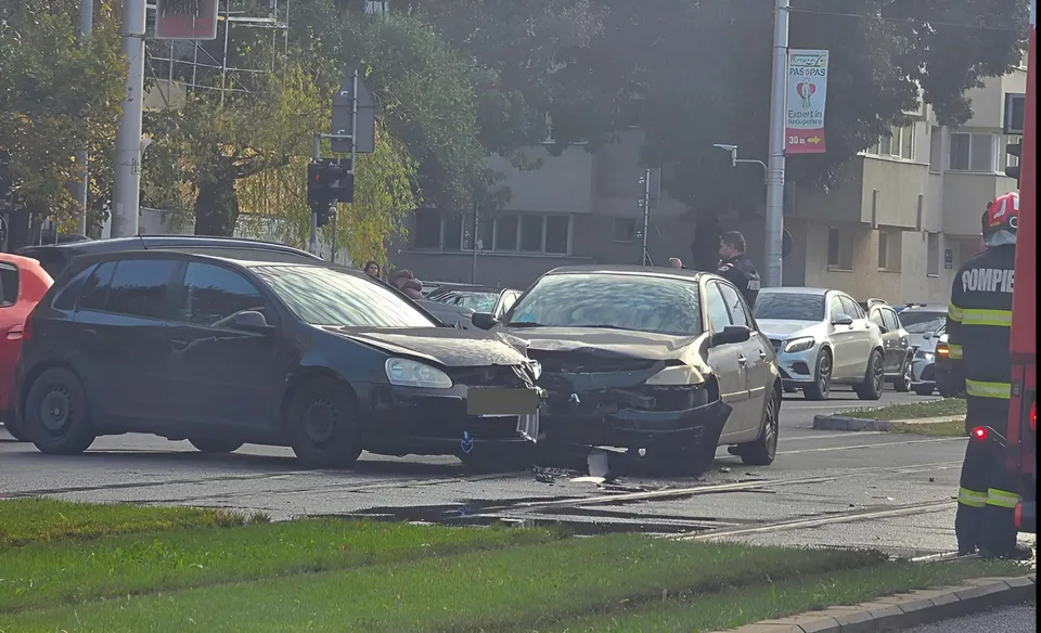 Două autoturisme de culoare închisă sunt oprite pe o stradă lângă o zonă cu iarbă, în prezența unui membru al corpului de pompieri.