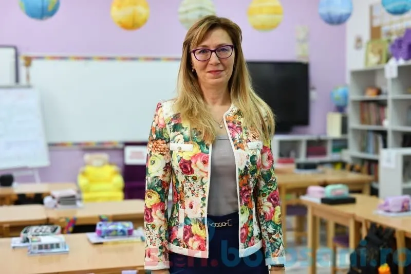 O femeie cu ochelari și o jachetă cu imprimeu floral stă în fața unor mese de clasă și rafturi cu cărți.