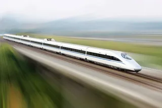 Un tren de mare viteză alb cu dungi albastre circulă pe o cale ferată într-un peisaj verde blurat de mișcare.