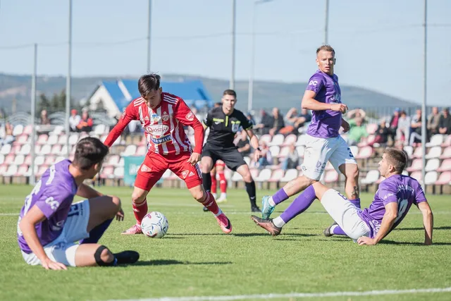 Cei patru jucători de fotbal de pe teren sunt îmbrăcați în echipamente sportive colorate, un jucător fiind în proces de a lovi mingea albă.