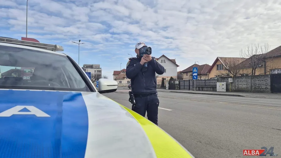 Un agent de poliție în uniformă întunecată ține un aparat de măsurat în mâini pe marginea unui drum asfaltat lângă o mașină albastră și albă.