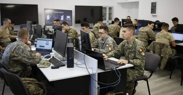 Soldați în uniforme de camuflaj stau la birouri cu computere și monitoare într-o încăpere cu pereți albi.