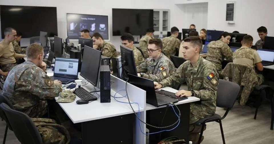 Soldați în uniforme de camuflaj stau la birouri cu computere și monitoare într-o încăpere cu pereți albi.