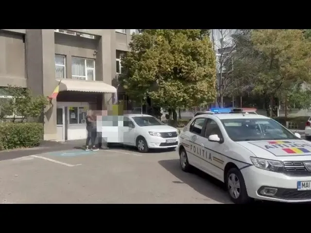 O mașină de poliție albă stă pe un drum lângă un vehicul alb și o clădire cu copaci în fundal.